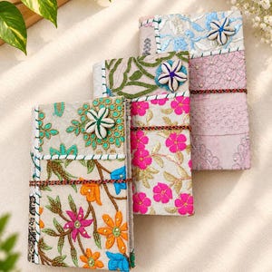 May include: Three rectangular journals featuring embroidered fabric covers with floral patterns in orange, pink, blue, and green. Each journal is secured with a thin, multi-coloured cord and decorated with a white, five-petalled shell flower accent.