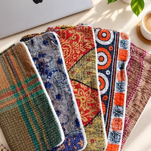 May include: A collection of five rectangular zippered pouches featuring various quilted fabric patterns, including green plaid, blue floral, red and yellow botanical, orange geometric circles, and a brown textured design with white stitching.
