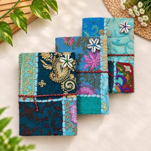 Peut inclure: Trois carnets rectangulaires dotés de couvertures en patchwork dans des tons bleu, sarcelle et vert. Chaque couverture présente des broderies complexes, des accents de fil métallique doré et une fleur en coquillage blanc attachée par un cordon.