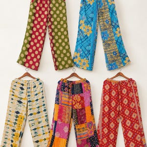 Peut inclure: Cinq paires de pantalons à jambes larges suspendus sur des cintres en bois. Les motifs incluent vert olive à pois rouges, bleu à fleurs jaunes, crème à motifs tie-dye bleu et jaune, un patchwork multicolore et rouge à motifs géométriques.