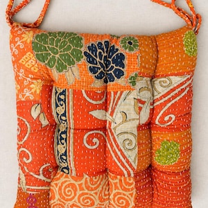 May include: A square seat cushion featuring a patchwork design of orange fabric with various floral and swirling patterns in green, navy blue, and cream. The cushion includes white hand-stitched detailing and two orange fabric ties at the top corners.