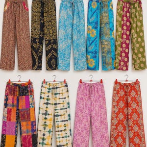 Könnte beinhalten: Eine Kollektion von neun Hosen mit weitem Bein, die an einzelnen Kleiderbügeln hängen. Die Hosen weisen verschiedene bunte Muster auf, darunter florale, geometrische, gepunktete, Batik- und Patchwork-Designs in Rot-, Blau-, Gelb-, Rosa- und Grüntönen.