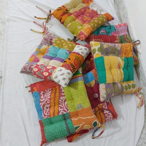 Könnte beinhalten: Eine Sammlung bunter Sitzkissen im Patchwork-Stil. Die Kissen weisen eine Vielzahl von Mustern und Farben auf, darunter Rot, Orange, Grün und Gelb. Jedes Kissen hat Bänder zur Befestigung an einem Stuhl. Die Kissen sind auf einer weißen Oberfläche angeordnet.