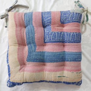 May include: Square cushion with a patchwork design in pink, blue, and cream. The cushion has a quilted texture and is tied with fabric straps. The design features stripes and patterned squares.