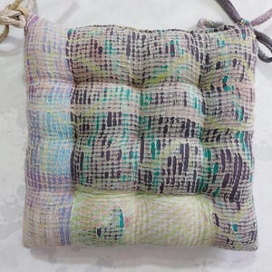 May include: Square seat cushion with a quilted design. The cushion features a patchwork pattern in shades of purple, blue, green, and yellow. It has ties at the corners for securing to a chair. The cushion is approximately 16 inches square.