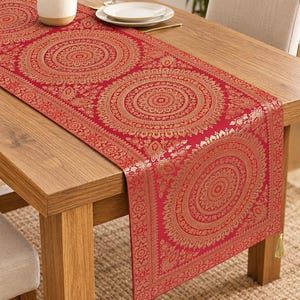 May include: A rectangular red table runner featuring a repeating gold circular mandala pattern and intricate floral borders. The fabric is finished with small, light-coloured tassels at each corner, draped over a wooden dining table surface.