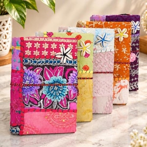 Könnte beinhalten: Eine Reihe von fünf rechteckigen Notizbüchern mit Patchwork-Stoffeinbänden in leuchtenden Farben wie Pink, Gelb, Hellblau, Orange und Lila. Jedes Notizbuch ist mit aufwendigen Stickereien, Pailletten und einem weißen Muschelakzent verziert.