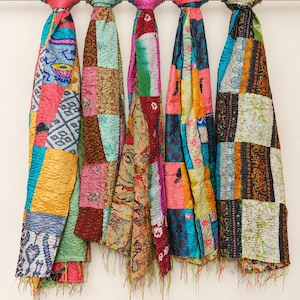 May include: Five rectangular scarves hanging from a horizontal metal rod. Each scarf features a patchwork design with various colorful fabric squares in shades of blue, pink, yellow, green, and brown, finished with frayed edges at the bottom.