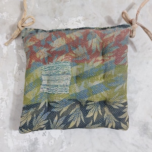 Könnte beinhalten: Quadratisches Kissen mit Patchwork-Design. Das Kissen weist eine Mischung aus Farben auf, darunter Rot, Blau, Grün und Marine. Es hat ein Blumenmuster am Boden und ist mit beigefarbenen Kordeln gebunden.