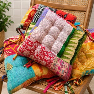 Puede incluir: Una pila de cojines cuadrados para asiento que presentan un mosaico de telas coloridas con varios patrones, incluyendo motivos florales, geométricos y animales. Cada cojín está acolchado con costuras visibles y tiene lazos de tela.
