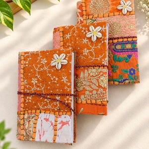 Könnte beinhalten: Drei rechteckige Notizbücher mit Patchwork-Einbänden in leuchtenden Orange-, Rosa- und Goldtönen. Jedes Notizbuch ist mit aufwendigen Stickereien, metallischen Fadenverzierungen und einer weißen Muschelblüte versehen und mit einer Kordel verschlossen.