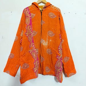 Puede incluir: Una chaqueta con capucha de color naranja brillante con cierre de cremallera frontal y mangas largas. La tela está decorada con un patrón de cachemira repetitivo en tonos rosa y dorado, con costuras blancas visibles en toda la prenda.