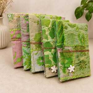 May include: A set of five rectangular journals featuring patchwork fabric covers in shades of green and pink. Each journal is decorated with intricate floral embroidery, beadwork, and a single white flower-shaped button closure on the front cover.