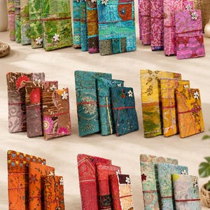 Könnte beinhalten: Eine Sammlung rechteckiger Stofftagebücher mit verschiedenen bunten Patchwork-Mustern, aufwendigen Stickereien und Ziernähten. Jedes Tagebuch ist mit einer dünnen roten Kordel verschlossen und mit einem kleinen weißen Muschelknopf verziert.