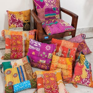 May include: A collection of square patchwork cushions featuring vibrant, multi-colored fabric panels with intricate kantha stitching. Each cushion displays a unique combination of floral, geometric, and abstract patterns in shades of pink, orange, yellow, and blue, with some cushions featuring corner ties.