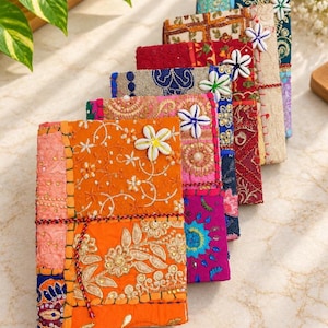 Könnte beinhalten: Eine Sammlung von sechs rechteckigen Notizbüchern mit Patchwork-Stoffeinbänden in leuchtenden Farben wie Orange, Pink, Rot, Blau und Gold. Jedes Notizbuch ist mit aufwendigen Stickereien, Metallic-Fäden und einer weißen Muschelblüte verziert.