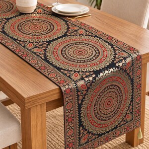 Puede incluir: Un camino de mesa rectangular largo con fondo negro y patrones circulares de mandala repetidos en rojo, dorado y crema. La tela tiene una textura tejida detallada con bordes florales ornamentados y borlas de color verde claro en los extremos.