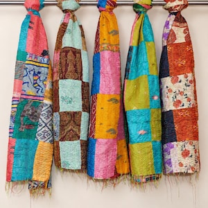 May include: Five rectangular fabric scarves hang vertically from a metal rod. Each scarf features a patchwork design of various colourful textiles with intricate patterns, including floral motifs, geometric shapes, and animal figures, finished with frayed edges.
