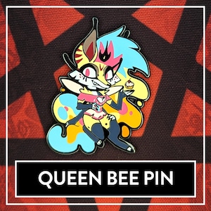 Könnte beinhalten: Eine farbenfrohe Emaille-Anstecknadel mit einer Cartoon-Figur mit gelbem, blauem und rosa Fell, die ein rosa Kleid trägt und ein Cupcake hält. Die Anstecknadel trägt unten den Schriftzug "Queen Bee Pin".
