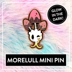 Könnte beinhalten: Eine goldfarbene Morelull-Mini-Anstecknadel mit einem weißen, zwinkernden Gesicht und drei rosa und violetten Pilzen. Die Anstecknadel leuchtet im Dunkeln.