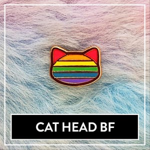 Könnte beinhalten: Ein goldfarbener Ansteckpin in Form eines Katzenkopfes mit Regenbogenmuster und roten Ohren. Der Pin hat den Text "CAT HEAD BF" in einem schwarzen Rechteck.