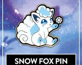 May include: A metal enamel pin featuring a stylized white fox with light blue accents, large blue eyes, and a fluffy tail, holding a small white snowflake icon. The pin is displayed above a black rectangular label with the text SNOW FOX PIN.
