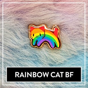 Könnte beinhalten: Eine goldfarbene Anstecknadel mit einem Cartoon-Katzen-Design mit einem Regenbogenkörper. Die Katze ist goldfarben umrandet und hat ein lächelndes Gesicht. Der Text "RAINBOW CAT BF" wird in einem schwarzen Rechteck darunter angezeigt.