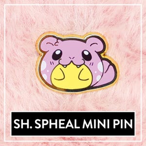 May include: A gold-colored enamel pin featuring a cartoon Spheal, a Pokemon, with a pink body, yellow belly, and white spots. The pin is on a pink fuzzy background. The text "SH.SPHEAL MINI PIN" is below the pin.