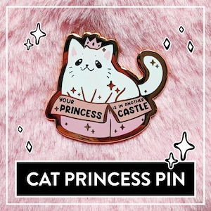 Puede incluir: Pin de esmalte dorado con un gato blanco con una corona, sentado en una caja con el texto "Your Princess is in another Castle".