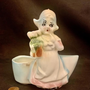 May include: A vintage ceramic planter shaped like a girl wearing a blue and white hat and dress. She is holding a bouquet of flowers and has a surprised expression on her face.