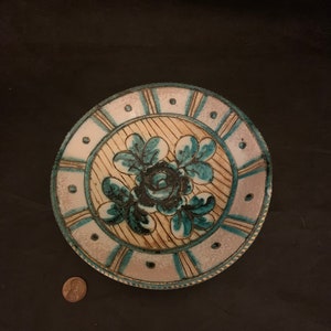 May include: A round ceramic plate with a white background and a blue and green floral design. The plate has a gold rim and a white and gold border.