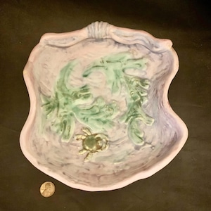 May include: A ceramic bowl with a pink, purple, and green glaze. The bowl is shaped like a seashell and features a crab and seaweed design.