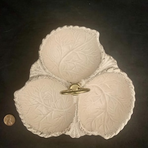 May include: A white ceramic serving dish with three sections shaped like cabbage leaves. The dish has a gold metal handle in the center.