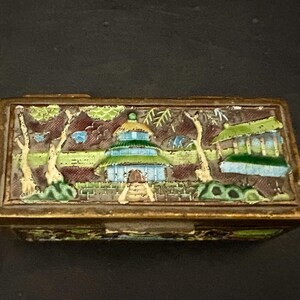 May include: A small, rectangular, brass box with a hinged lid. The lid is decorated with a colorful enamel scene of a pagoda in a garden setting. The scene features trees, flowers, and a path leading to the pagoda.