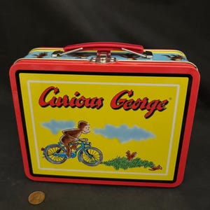 May include: Red metal lunchbox with a handle. The front of the lunchbox features a yellow background with the words "Curious George" in red and black lettering. A cartoon image of Curious George riding a blue bicycle is in the center of the lunchbox.