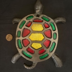 May include: A dark gray metal turtle wall hanging featuring a shell with translucent red, yellow, and green glass inserts. The turtle has a textured head, legs, and tail. A copper-colored penny is placed to the left for scale reference.