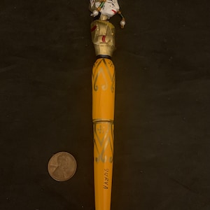 May include: A wooden pen with a carved figure of a person wearing a red hat and gold clothing. The pen is painted yellow with gold accents and the word "SURYA" is written on the bottom.