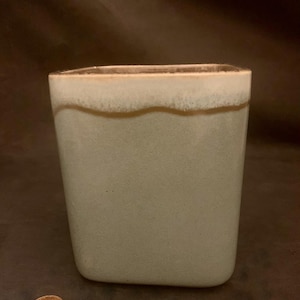 May include: A light green ceramic vase with a brown rim and a white glaze. The vase has a rectangular shape and a slightly rounded bottom.