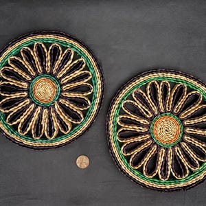 May include: Two woven trivets with a black, green, and brown daisy design. The trivets are round and have a raised center.