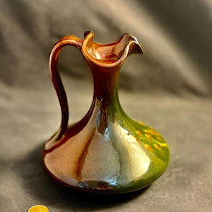 May include: A ceramic pitcher with a brown handle and rim, transitioning to a green body. The pitcher has a glossy finish and a unique, ruffled rim. A gold coin is in the foreground.