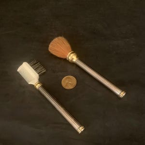 May include: A vintage beauty set featuring a small comb and a makeup brush. Both have silver-toned, ribbed handles with gold accents. The brush has a soft, brown bristle head. A penny is included for size reference.