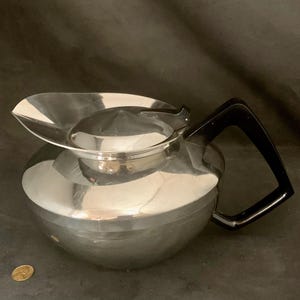 May include: A silver coffee pot with a black handle. The pot has a rounded body and a wide, flat lid.