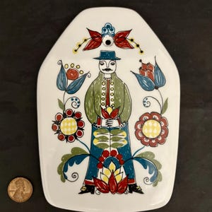May include: A white ceramic tile with a folk art design featuring a man in a green jacket, blue pants, and a black hat. The man is surrounded by red, yellow, and blue flowers.