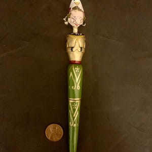 May include: A decorative pen with a painted wooden design. The pen features a green barrel with gold accents and a detailed figure at the top. A penny is next to the pen for scale.