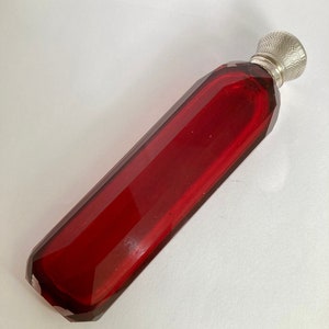 May include: A red glass bottle with a silver stopper. The bottle is faceted and has a long, slender shape.