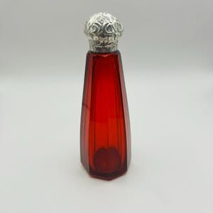 May include: A tall, faceted ruby red glass bottle with a tapered, hexagonal body. The bottle is topped with a decorative, rounded silver-colored metal cap featuring intricate embossed floral and scrollwork patterns across its entire surface.