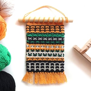 Puede incluir: Un tapiz tejido con un patrón geométrico en naranja, negro, blanco y verde. El textil está sujeto a un palillo de madera con una cuerda para colgar y tiene flecos naranjas. Ovillos de lana en naranja, verde y negro en el fondo.