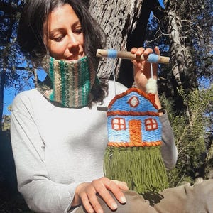 May include: A woven wall hanging in the shape of a house, featuring a blue roof, orange door and windows, and green fringe. The house is suspended from a wooden dowel with blue yarn accents. The overall design is handmade and rustic.