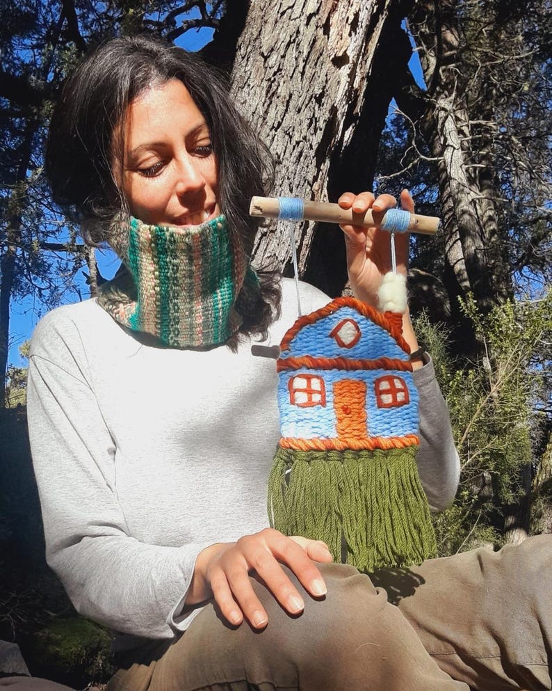 May include: A woven wall hanging in the shape of a house, featuring a blue roof, orange door and windows, and green fringe. The house is suspended from a wooden dowel with blue yarn accents. The overall design is handmade and rustic.