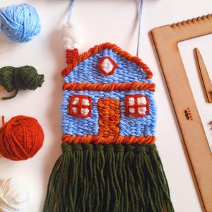 May include: A handmade woven wall hanging in the shape of a house. The house is blue with orange trim, a white chimney, and green fringe. Blue yarn is used for the hanging cord. Additional yarn balls in blue, orange, green, and white are scattered around the house.